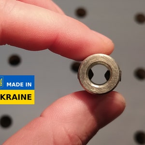 May include: A small, metallic, cylindrical object with a central aperture and a star-shaped interior. The object is held between fingers, with a blurred background. The image includes a "MADE IN UKRAINE" label.