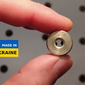 Puede incluir: Primer plano de un anillo de metal plateado con un pequeño agujero en el centro. El anillo está siendo sostenido por una mano. El fondo está desenfocado y muestra una superficie blanca con puntos negros.