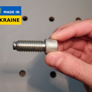 Könnte beinhalten: Eine silberfarbene Schraube mit gerändeltem Kopf und Gewindeschaft. Die Schraube wird in einer Hand gehalten. Das Bild enthält ein blaues und gelbes Rechteck mit dem Text "MADE IN UKRAINE".