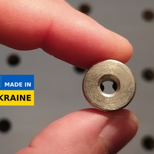 May include: A small, metallic, circular object with a central hole and a star-shaped interior. The object is held between fingers. A blue and yellow label with the text "MADE IN UKRAINE" is visible.