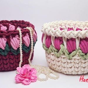 Puede incluir: Dos cestas de ganchillo hechas a mano con un diseño floral. Una es de color burdeos con detalles en rosa y verde, la otra es de color blanco roto con detalles en rosa y verde. Un collar de perlas y una flor rosa están junto a las cestas. El texto "Haekelqueenart" está en la esquina inferior derecha.