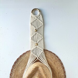 Peut inclure: Un porte-chapeau en macramé blanc avec des perles dorées suspendu à un mur blanc avec un chapeau de paille suspendu.