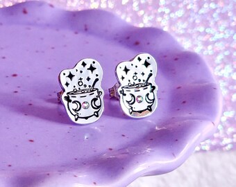 May include: A pair of silver earrings featuring a cauldron with stars and moons, accented with pink glitter. The earrings are displayed on a purple cake stand.