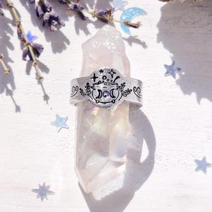 May include: A silver ring with a detailed design of a cauldron with stars, moons, and herbs. The ring is on a white crystal.