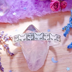 May include: A white headband with a repeating black cat face design is displayed on a clear crystal. The headband is surrounded by lavender, dried roses, and a bundle of sage. The background is a fluffy purple surface with decorative stars and moons.