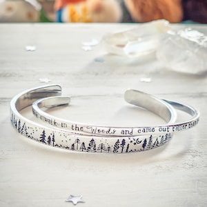 May include: Two silver-colored metal bracelets with an open cuff design. One bracelet features a forest silhouette with stars and a crescent moon. The other bracelet has the text: "I took a walk in the Woods and came out taller than trees."