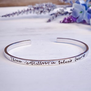 May include: Silver bangle bracelet with the inscription "Throw glitter in today's face" with two stars on either side of the word glitter.