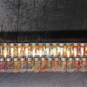 May include: Rows of small glass bottles with silver lids, filled with colorful liquids and decorative elements. The bottles are arranged in a semi-circular pattern against a dark background, creating a visually appealing display.