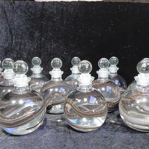 May include: A collection of clear glass bottles with round bodies and clear glass stoppers. Each bottle has a brown and gray design around the middle. The bottles are arranged on a dark surface.