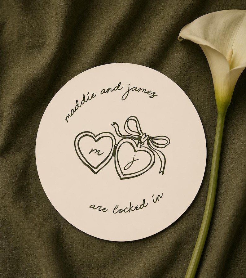 Pu&ograve; includere: Un sottobicchiere rotondo color crema con il testo "maddie and james" e "are locked in". Due medaglioni a forma di cuore sono collegati al centro, ognuno con una lettera. Un giglio bianco sullo sfondo.
