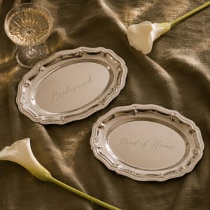 Op de afbeelding: Twee ovale zilverkleurige dienbladen met geschulpte randen. Eén dienblad is gegraveerd met het woord Bridesmaid in cursief schrift, en het andere dienblad is gegraveerd met de woorden Maid of Honour in bijpassend cursief schrift. Beide dienbladen zijn reflecterend.