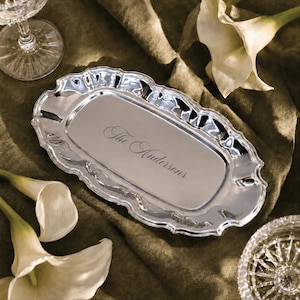 May include: An oval silver-toned metal tray with a scalloped edge and a polished reflective surface. The centre of the tray features the engraved script text "The Andersons" in a cursive font, centred horizontally on the smooth metal base.