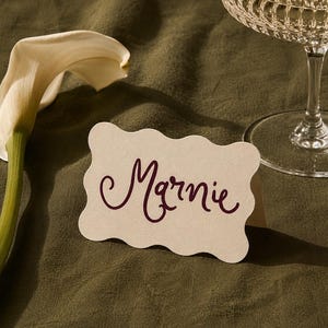 May include: A cream-coloured, scalloped-edge place card featuring the name Marnie written in a dark purple, cursive script. The card is positioned next to a white calla lily flower and the base of a clear, textured glass goblet.