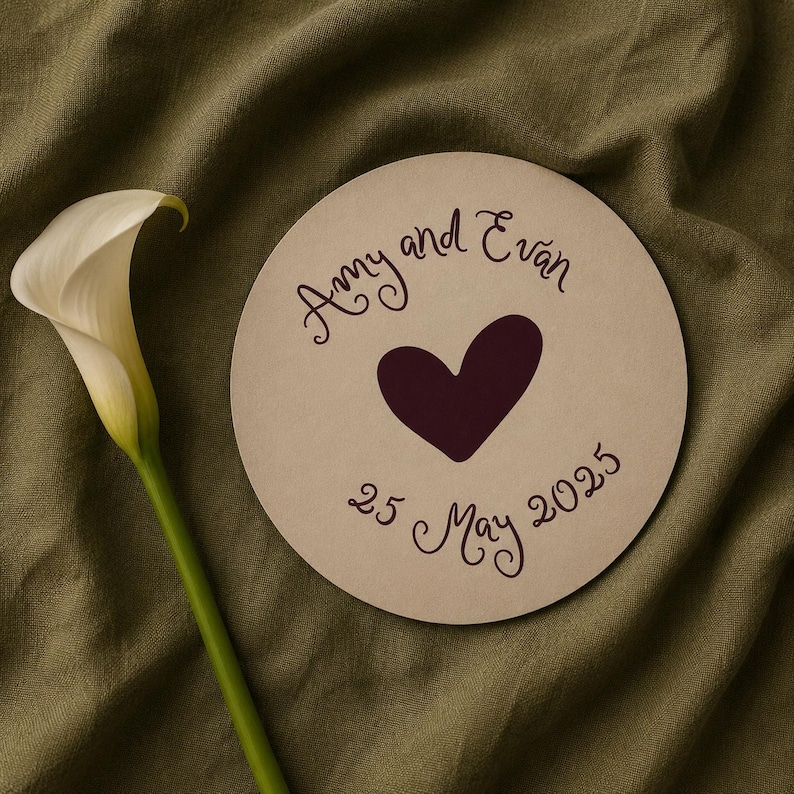 May include: A beige circular wedding announcement with the names "Amy and Evan", a dark purple heart, and the date "25 May 2025". A white calla lily with a green stem is on the left.
