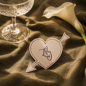 May include: A heart-shaped paper card featuring a dark brown outline of an arrow piercing through it. The name Lily is written in the center in a dark brown cursive script. The card is positioned near a clear glass goblet and a white calla lily.