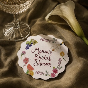 Può includere: Un sottobicchiere circolare in carta con bordo smerlato che riporta la scritta Maria's Bridal Shower in corsivo rosso scuro. Il design include illustrazioni ad acquerello di uva viola, fiori rosa, una fetta di pompelmo e rametti verdi.