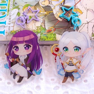 Puede incluir: Dos charms de acrílico de personajes de anime. El charm de la izquierda presenta a una chica con cabello morado y un vestido blanco. El charm de la derecha presenta a una chica con cabello blanco y un vestido blanco. Ambos charms tienen un diseño de estrella azul.