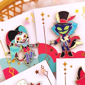 May include: Two enamel pins featuring characters from the animated series Helluva Boss. The first pin is of a jester with a red, black, and blue costume. The second pin is of a blue-haired demon with a red and white striped suit and a top hat.