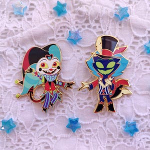 May include: Two enamel pins featuring cartoon characters. The first pin is a blue, red, and yellow jester with a wide smile. The second pin is a green and blue cat-like character wearing a red and white hat and a red and white striped shirt.