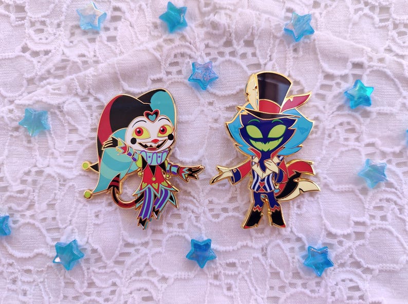 May include: Two enamel pins featuring cartoon characters. The first pin is a blue, red, and yellow jester with a wide smile. The second pin is a green and blue cat-like character wearing a red and white hat and a red and white striped shirt.