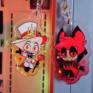 Peut inclure: Deux porte-clés en acrylique représentant des personnages de dessins animés. L'un présente un personnage en costume blanc et haut de forme, tenant une pomme. L'autre présente un personnage rouge avec une cape. Les deux ont des attaches à chaîne argentée.