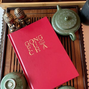 May include: A red book with gold lettering reading "GONG FU CHA" sits on a bamboo tea tray, surrounded by tea cups, a teapot, and decorative figurines. The tea set includes a green teapot and two matching cups.
