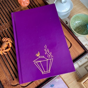 Può includere: Un diario viola con un designo dorato di una teiera con vapore che esce dal beccuccio e una farfalla che vola sopra. Il testo "Gong Fu Cha" è scritto in un carattere stilizzato.