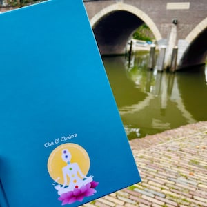 May include: A teal blue journal with a sticker of a person sitting in a lotus position with seven colored chakras. The text "Cha & Chakra" is printed on the journal.