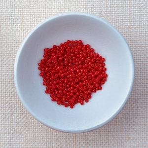 May include: A white ceramic bowl filled with a pile of small, vibrant red beads. The beads are densely packed, creating a textured surface. The bowl has a simple, rounded shape and sits on a neutral-toned fabric background.