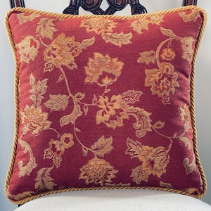 May include: Square decorative pillow with a burgundy background and a floral pattern in shades of gold and tan. The pillow has a gold rope trim.