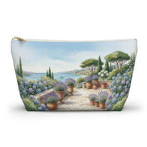 Puede incluir: Un neceser con cremallera con una pintura de acuarela de una terraza costera. La obra de arte representa plantas de lavanda en macetas de terracota, hortensias azules y una vista del mar bajo un cielo azul. El neceser tiene una parte superior crema y una cremallera dorada.