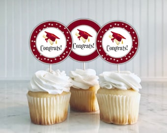 Pode incluir: Três cupcakes cobertos com glacê branco, cada um adornado com um topper temático de formatura. Os toppers apresentam um capelo vermelho, um diploma e a palavra "Congrats!" em uma fonte elegante, perfeito para uma celebração de formatura.