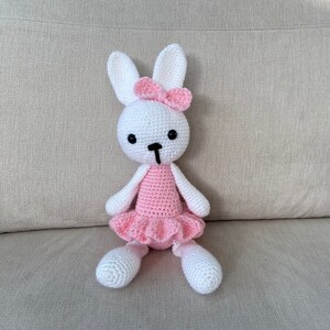 Puede incluir: Un conejito de crochet blanco con un tutú rosa y un lazo rosa. El conejito está sentado en un sofá marrón claro.