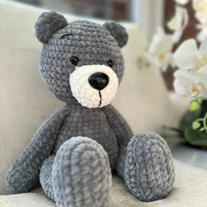 May include: A grey crocheted teddy bear with white accents around the nose and mouth. The bear is sitting on a white surface.