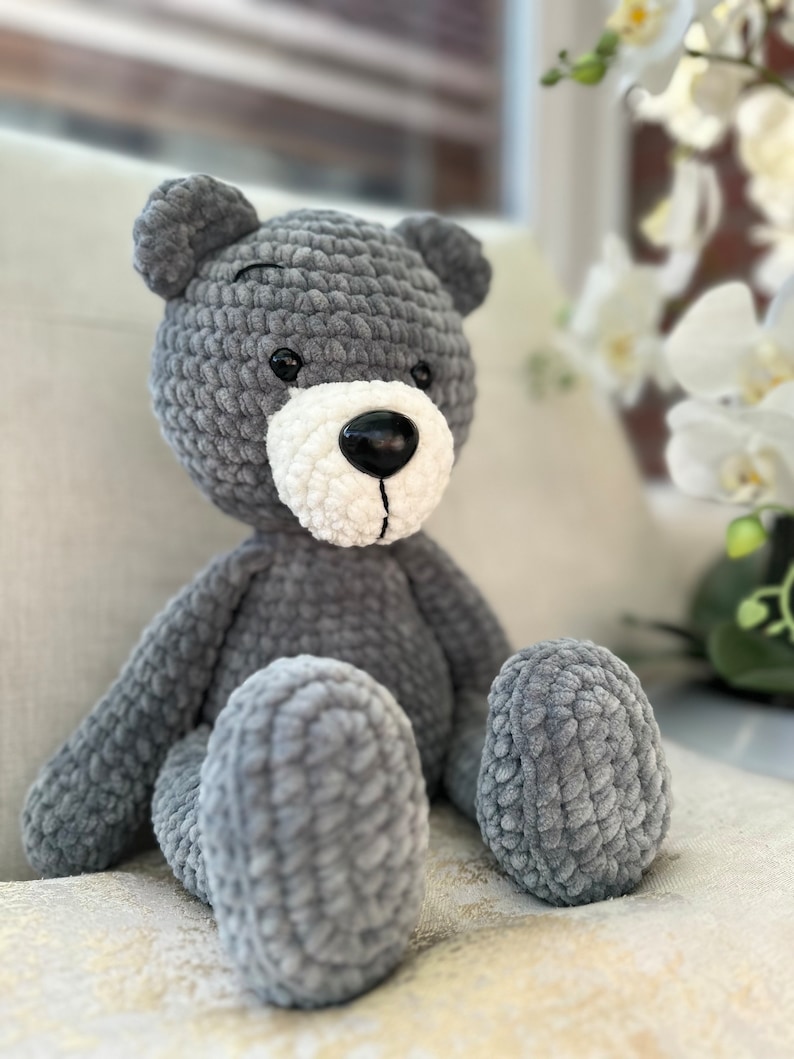 K&ouml;nnte beinhalten: Ein grauer geh&auml;kelter Teddyb&auml;r mit wei&szlig;en Akzenten um Nase und Mund. Der B&auml;r sitzt auf einer wei&szlig;en Oberfl&auml;che.