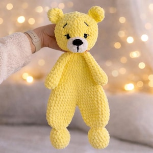 May include: A handmade yellow crochet bear doll with a black nose and small black eyes. The bear has a red collar and small ears. The doll is being held up in front of a blurred background of lights.