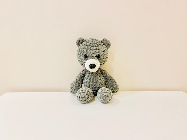Puede incluir: Un oso de peluche gris tejido a crochet con detalles en fieltro blanco para los ojos y la nariz. El oso est&aacute; sentado sobre una superficie blanca.