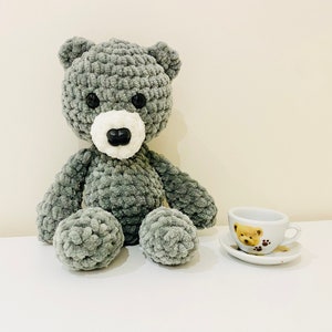 Puede incluir: Un oso de peluche gris tejido a crochet con ojos negros y una nariz blanca se sienta sobre una superficie blanca junto a una peque&ntilde;a taza de t&eacute; blanca con un dise&ntilde;o de oso marr&oacute;n.