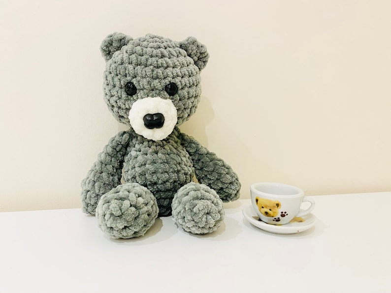 Puede incluir: Un oso de peluche gris tejido a crochet con ojos negros y una nariz blanca se sienta sobre una superficie blanca junto a una peque&ntilde;a taza de t&eacute; blanca con un dise&ntilde;o de oso marr&oacute;n.