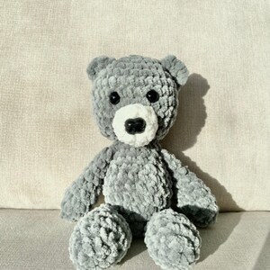 Puede incluir: Un oso de peluche gris tejido a crochet con detalles blancos alrededor de la nariz y la boca. El oso est&aacute; sentado sobre una superficie texturizada gris claro.