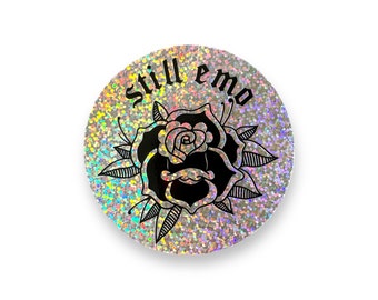 May include: A round, iridescent sticker featuring a black and white rose with the text "still emo" in a black gothic font.