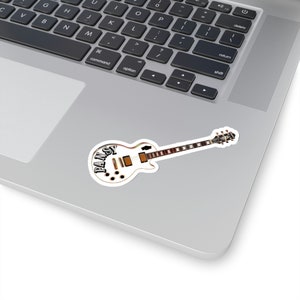 Pode incluir: Uma guitarra el&eacute;trica branca com um design preto e marrom. A guitarra tem a palavra "PANSY" escrita no corpo.