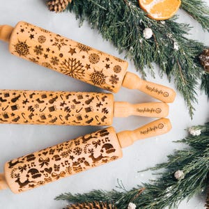 May include: Three wooden rolling pins with engraved holiday designs. One pin features snowflakes, another has Christmas-themed icons, and the third displays reindeer and gifts. Each pin has a wooden handle with the text "HappyRollingPin".