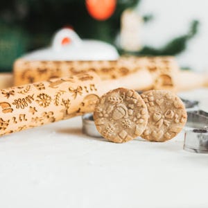 Può includere: Due biscotti di pan di zenzero con un design natalizio, realizzati con un mattarello con un design simile. I biscotti sono su una superficie bianca accanto a uno stampino per biscotti in metallo.