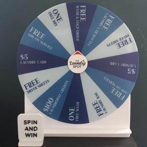 May include: A white and blue prize wheel with various laundry-related offers. The wheel features text such as "FREE Dryer Sheets", "$5 Laundry Card", and "Free Add-on Service". A small sign reads "Spin and Win".