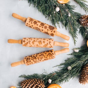 May include: Three wooden rolling pins with engraved holiday designs, including snowflakes, stars, and Christmas-themed icons. The rolling pins have light brown handles and are arranged on a light blue surface with evergreen branches and pine cones.