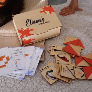 May include: A wooden game box with the text "Elison's GAME BOX" and red and orange abstract designs. The box is open, revealing game pieces, cards, and wooden tiles with bird illustrations. A hand is visible in the background.