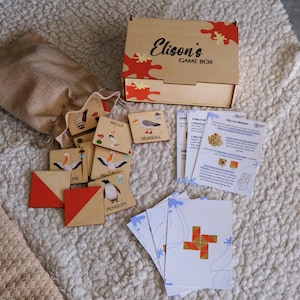 May include: A wooden game box with the text "Elison's GAME BOX" and red and orange accents. The box is open, revealing game pieces with bird illustrations and text. A burlap bag and cards are also present.