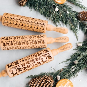 Puede incluir: Tres rodillos de madera con diseños navideños grabados. Uno presenta renos y estrellas, otro renos y árboles de Navidad, y el tercero renos, lazos y notas musicales. Los mangos son de madera clara, rodeados de vegetación.