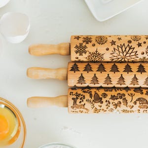 Könnte beinhalten: Drei hölzerne Nudelrollen mit eingravierten Feiertagsmotiven. Die obere Nudelrolle zeigt Schneeflocken und Sterne, die mittlere Nudelrolle hat Weihnachtsbäume und die untere Nudelrolle zeigt festliche Bilder. Die Nudelrollen haben hellbraune Griffe.